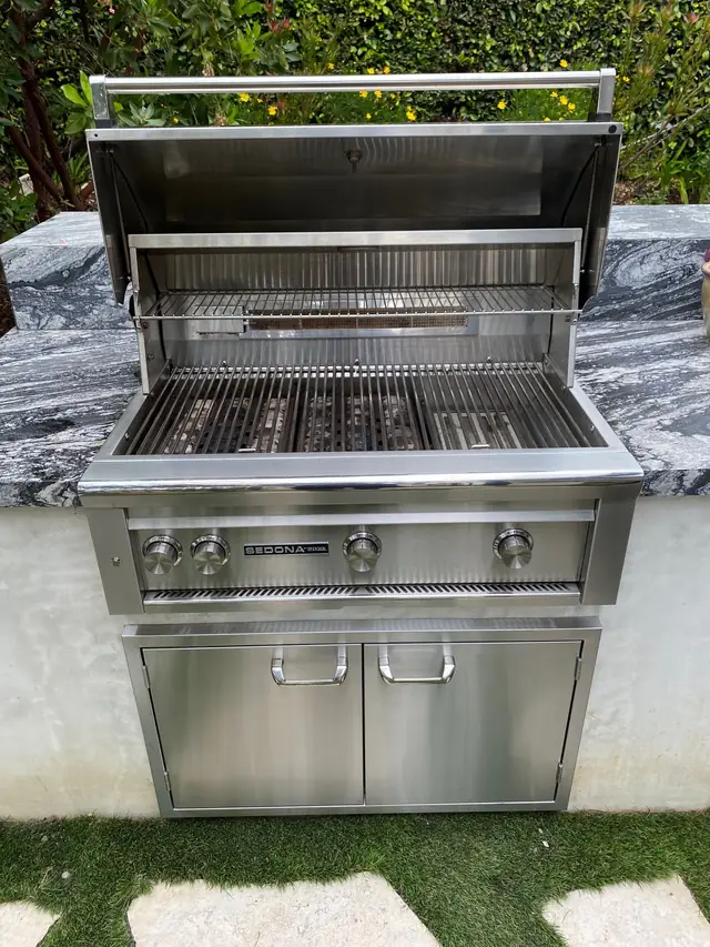 A clean stainless grill with its hood open above stainless cabinet doors on a stone counter