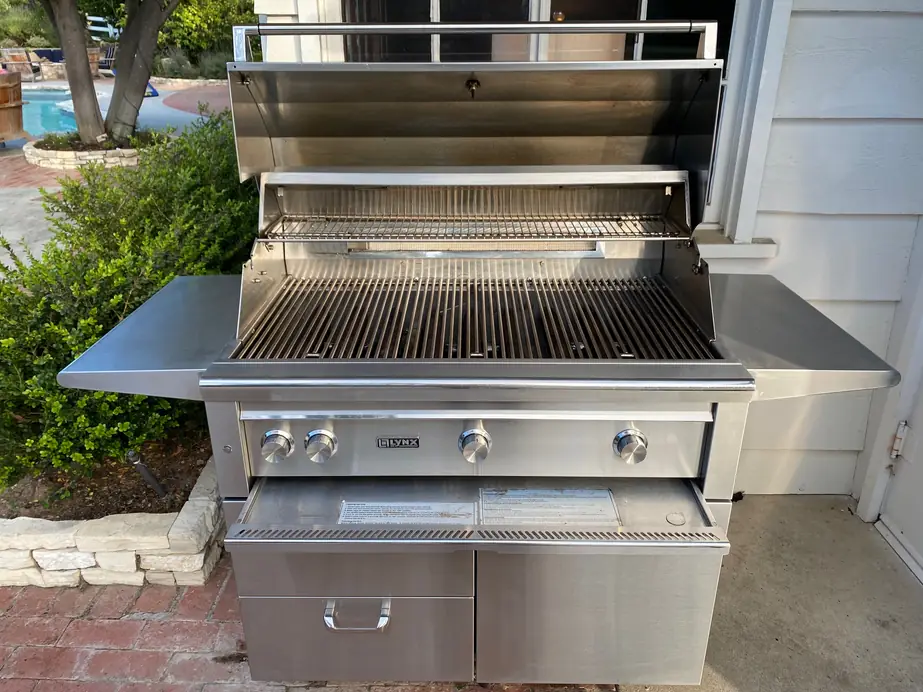 A freestanding stainless grill with its hood open beside a house and pool