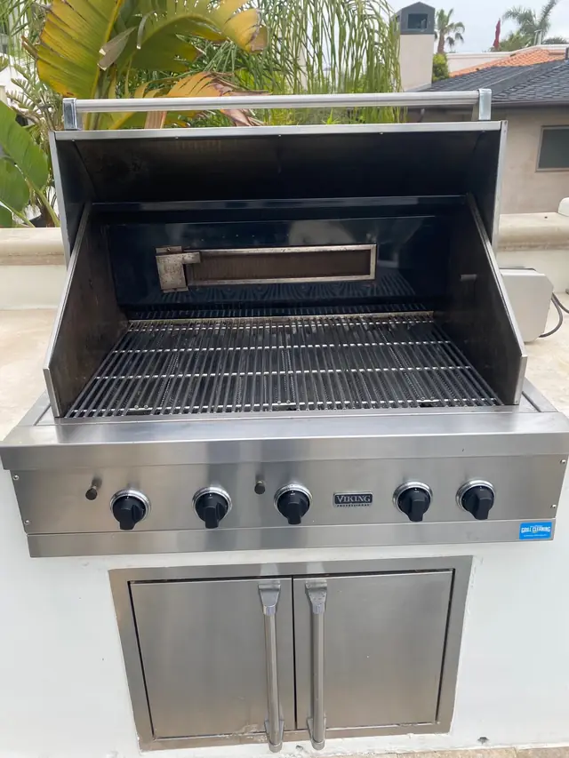 A clean built-in stainless grill with black knobs, palms behind it