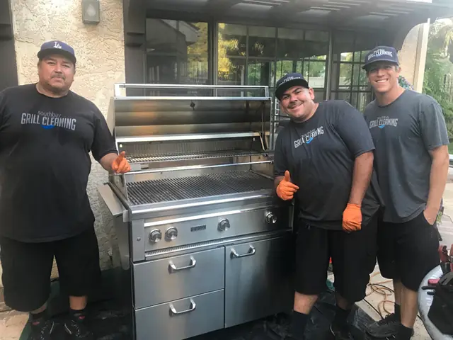 Three crew members in branded shirts and caps beside a finished grill